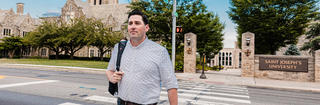 Doctoral student David Lloyd walks across Lapsley Lane on campus wearing a button up shirt and carrying a backpack