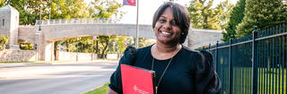 Charisse McGill '20 (MBA) stands on campus wearing a black blouse and carrying a red notebook