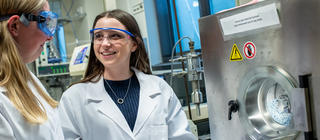 A young female student wearing a lab coat and goggles smiles at her colleague in a lab