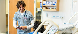 Male nursing student assisting female simulated patient in a hospital bed in the simulation lab