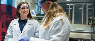 A young female student wearing a lab coat and goggles smiles at her colleague in a lab