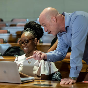 In a classroom, a professor and graduate student discuss work on the student's laptop