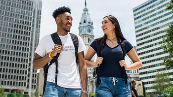 A male and female student are walking in center city Philadelphia with City Hall behind them in the distance